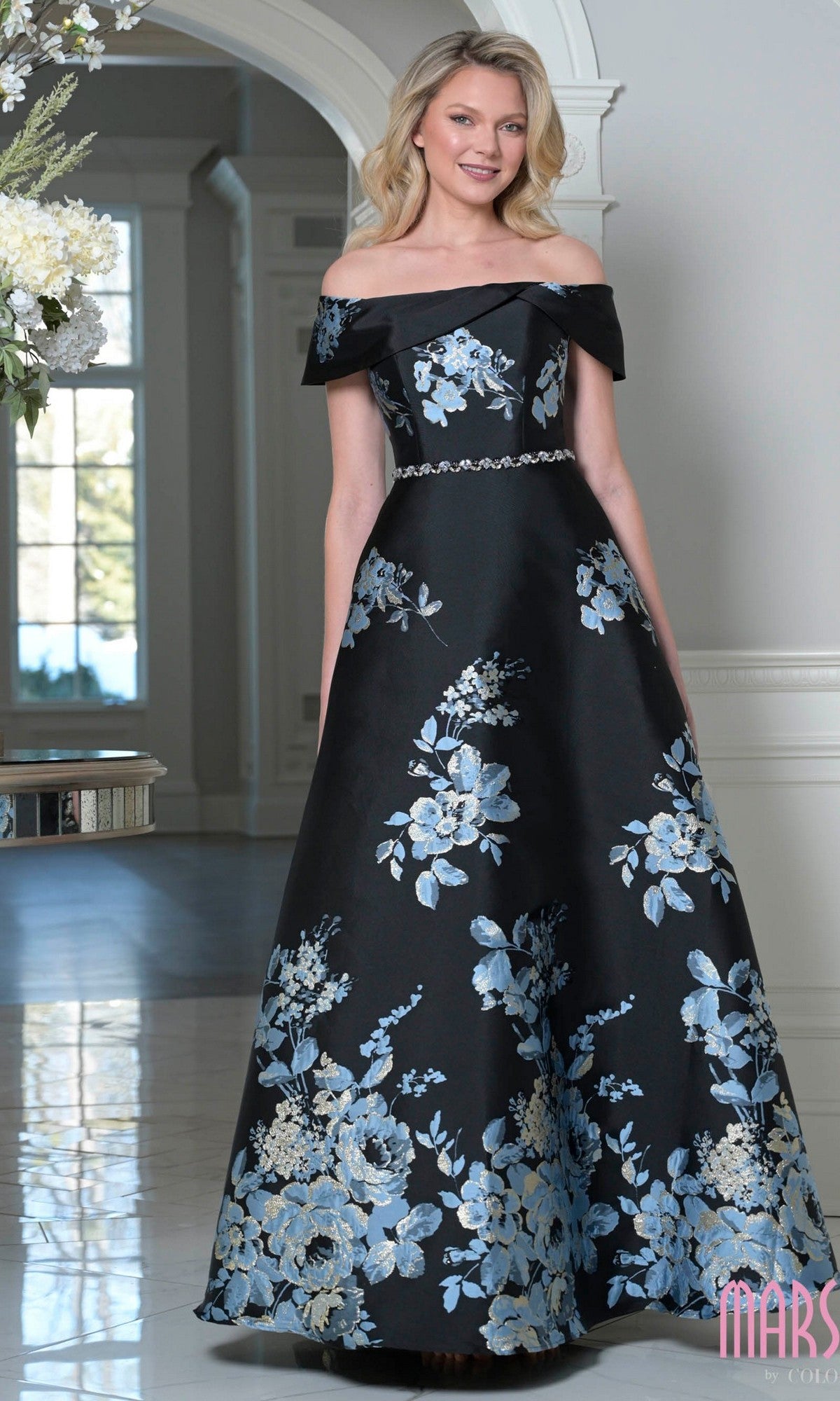 A woman wears the Marsoni MV1402 long formal dress in Black Multi, featuring an off-the-shoulder floral A-line design, while standing in a bright, elegant room with white walls and flowers nearby.